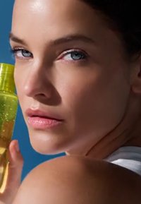 Primer plano de una mujer con ojos azules que sostiene una botella amarilla translúcida cerca de su rostro sobre un fondo azul.