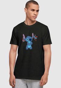 Camiseta negra de manga corta con un diseño de personaje de dibujos animados en azul. El personaje tiene orejas grandes y sostiene objetos rojos en cada mano.