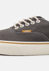Harmaa kangastennari beige-nauhoilla ja valkoisilla aukkoilla. Varusteena valkoinen kumipohja, jossa on tekstuuripintoja, sekä oranssi Vans-logotagi.