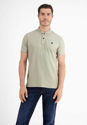 Mann trägt ein hellgrünes Poloshirt und dunkelblaue Jeans, steht mit neutralem Gesichtsausdruck vor weißem Hintergrund.