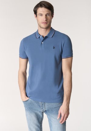 Uomo con polo blu con rifiniture rosa e jeans azzurri chiari, in piedi davanti a uno sfondo chiaro semplice.