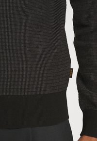 Maglione nero testurizzato con polsini e orlo a coste, caratterizzato da un motivo sottile. Include un piccolo etichetta arancione del marchio sul lato.