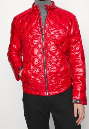 Homme portant une veste matelassée rouge brillante avec des coutures géométriques, une fermeture éclair noire et un col à boutons-pression sur un col roulé gris.