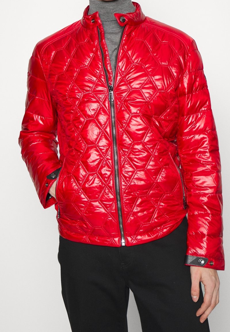Homme portant une veste matelassée rouge brillante avec des coutures géométriques, une fermeture éclair noire et un col à boutons-pression sur un col roulé gris.