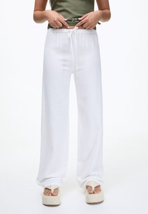 Woman wearing loose white drawstring pants, olive green top, and platform sandals, holding belt buckle at waist level.