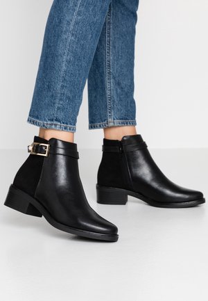 Sorte læderankelstøvler med sidelås og dekorativt guldspænde båret med blå beskårne jeans på hvidt gulv.