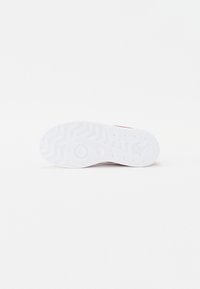 Hvit sneakersåle med et teksturert mønster, som har en sirkulær innrykning og fleksibelt, gummiaktig materiale. Røde detaljer er synlige på overdelen.
