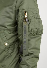 Veste bomber verte détaillée d'une fermeture éclair dorée, d'une poche latérale et d'une étiquette indiquant "BEFORE FLIGHT". Fabriquée en matériau lisse et matelassé.