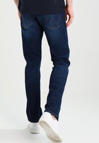 Vaqueros de mezclilla azul oscuro con un corte ajustado, que presentan bolsillos traseros con doble costura, un ligero desgaste y una textura suave. Llevados con zapatillas blancas.