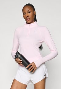 Top roz deschis cu mânecă lungă, cu fermoar jumătate și găuri pentru degete, asortat cu pantaloni scurți albi cu pliuri și o sticlă de apă neagră.