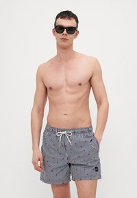 Bañador a rayas en azul marino y blanco con puntos rojos, cintura elástica, cordón ajustable y bolsillos laterales. Se llevan gafas de sol.