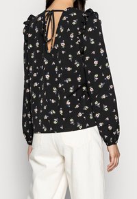 Blusa nera con motivo floreale, maniche a volant e design con apertura a chiave sul retro. Realizzata in tessuto leggero con polsini elastici.