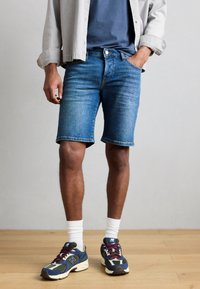 Τζιν σορτς σε ανοιχτό μπλε χρώμα, μήκους έως τη μέση του μηρού με κλασική ίσια γραμμή, φινιρισμένα με σκισμένη άκρη. Φορεμένα με πολύχρωμα αθλητικά παπούτσια.