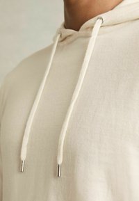Cremefarbener Kapuzenpullover mit weicher Textur, ausgestattet mit einem Kordelzug mit metallischen Spitzen und minimalistischer Gestaltung.