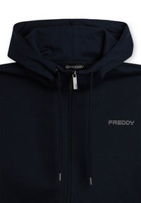 Sudadera con cremallera azul marino con cuello alto, cordones y herrajes plateados. Presenta un pequeño logo gris en el pecho izquierdo. Textura suave.