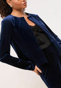 Tamnoplav kaput od baršuna s okruglim izrezom, dugim rukavima i skraćenim dizajnom. Crna čipkasta majica ispod dodaje teksturu i kontrast.