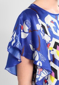 Silketop med blå bund, blomstermønster i hvid og rød, flæser i korte ærmer og geometriske accenter i sort, pink og gul.