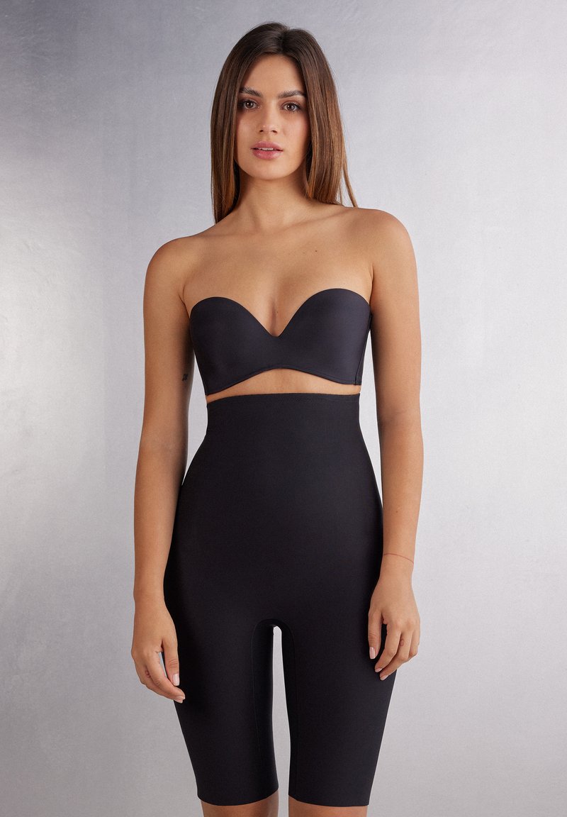 Zwarte strapless bodysuit van glad, rekbaar materiaal; heeft een uitsnede onder de borst en shortsachtige pijpen die tot halverwege de bovenbenen reiken.