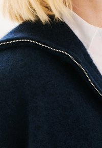 Granatowa wełniana kurtka o miękkiej teksturze, z metalowym suwakiem i lekko podniesionym kołnierzykiem. Pod kurtką widoczna biała koszula.