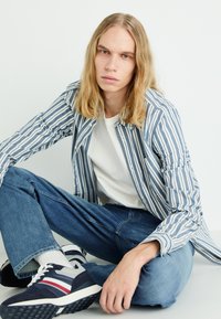 Camisa riscada azul e branca sobre uma t-shirt branca, acompanhada de jeans azuis e ténis marinhos com acentos em vermelho e branco. Roupa casual.