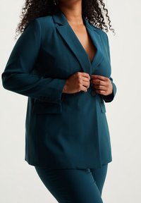 Tealblå skräddarsydd blazer med en enda knapplåsning, hakkragar, sidfickor och en slät textur, matchad med tealblå byxor.