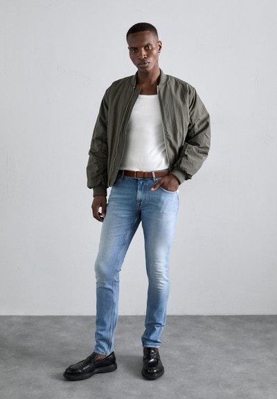 Chaqueta bomber caqui sobre una camiseta blanca sin mangas, combinada con jeans azul claro y zapatos negros. Texturas suaves, diseño ajustado y estilo casual.