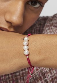 Pulsera de cordón rosa que presenta cuatro perlas blancas irregulares, con acentos plateados y un mecanismo de ajuste por nudo. Textura suave.