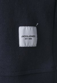Étiquette rectangulaire en tissu blanc avec texte noir "JACK & JONES EST. 1990" cousue sur un vêtement bleu marine foncé.