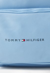 Pochette en tissu bleu clair avec une surface texturée, arborant un logo noir portant l'inscription "TOMMY HILFIGER" et un petit accent rouge et blanc.