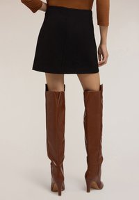 Minifalda de ante negra combinada con botas de tacón altas hasta la rodilla en color marrón, con un acabado suave y puntas afiladas, vista desde atrás.