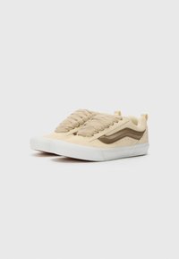 Beige suede sneakers with textured laces, brown accents, a white rubber sole, and distinctive side stripes. Short heel tab for easy wear.