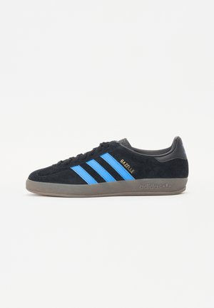 Czarny but sportowy Adidas Gazelle z niebieskimi paskami i brązową podeszwą, widok z boku na białym tle.