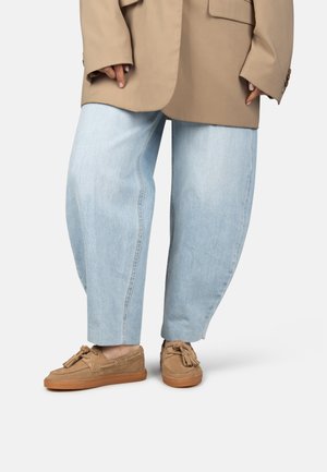 Persona que lleva mocasines beige con borlas, vaqueros de pierna ancha azul claro y una chaqueta beige extragrande, de pie frente a un fondo blanco.