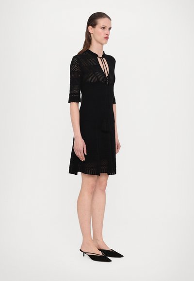 Schwarzes Strickkleid mit kurzen Ärmeln, Schlüssellochausschnitt, Spitzendetails und ausgestelltem Rock. Getragen mit schwarzen Absatzmules.