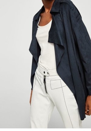 Femme portant une veste bleu marine ouverte sur le devant, un débardeur blanc et un pantalon blanc taille haute avec des coutures noires visibles et une fermeture éclair à l'avant.