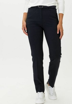 Vrouw met strakke zwarte broek met riem en witte sneakers, staand tegen een effen witte achtergrond.