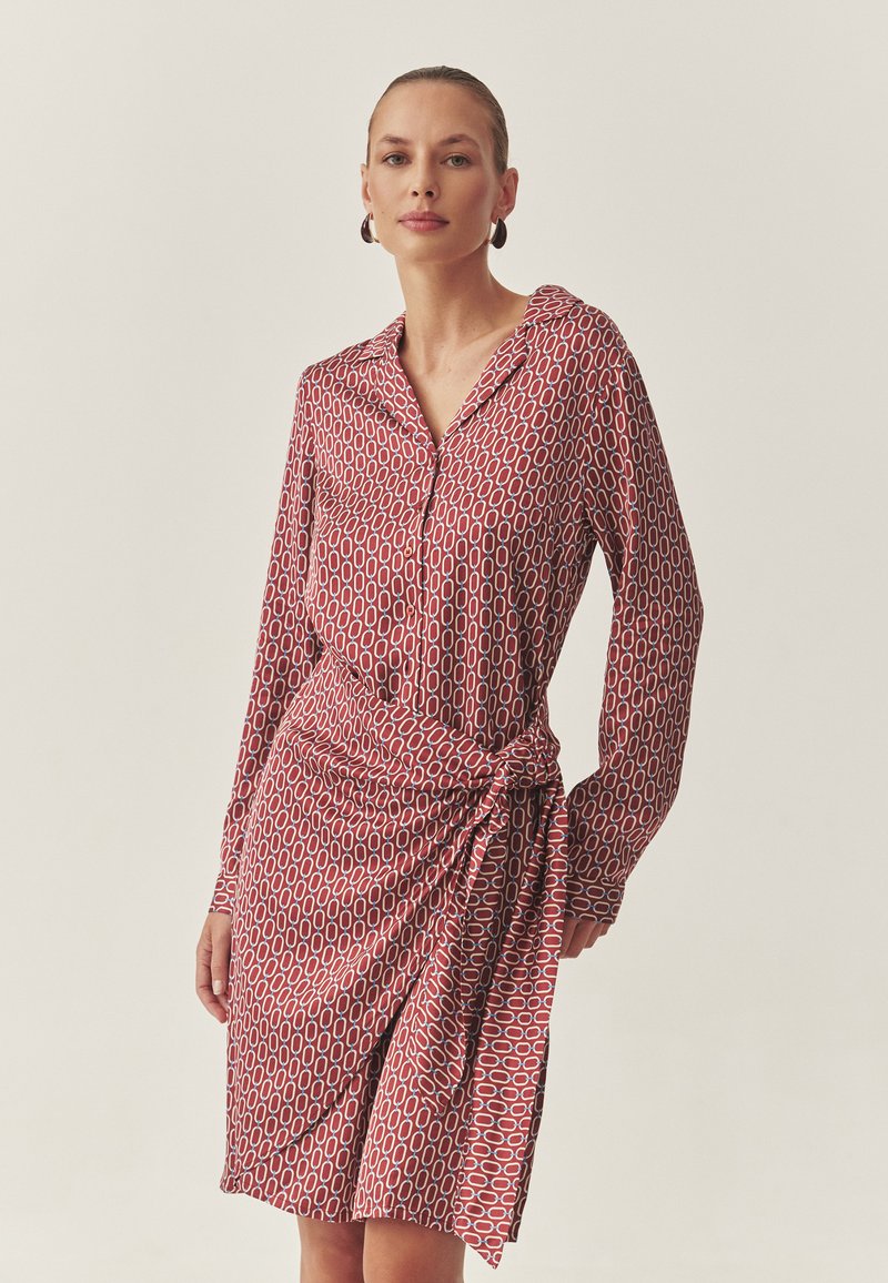 Rochie roșie și albă cu model, cu mâneci lungi, guler și detaliu legat în talie. Material neted, cu un luciu subtil, având un design geometric.