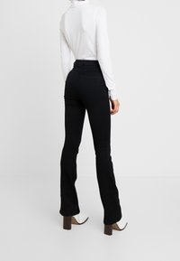 Personne portant un pantalon noir taille haute évasé, un col roulé blanc à manches longues et des bottines marron à talons blocs, debout devant un fond uni.