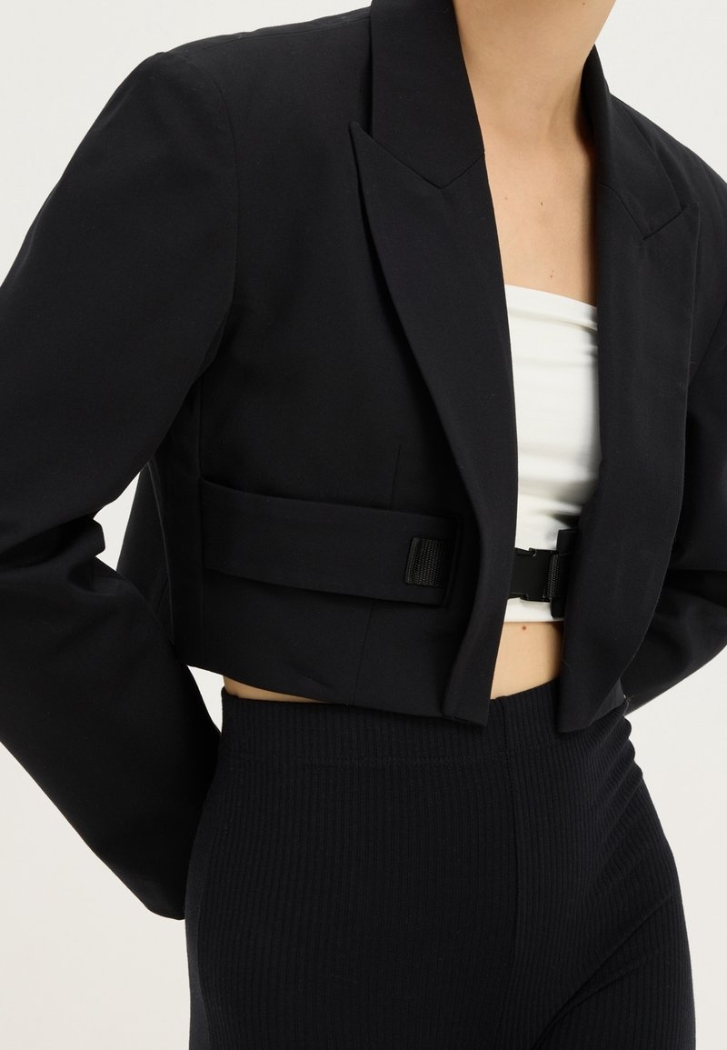 Personne portant un blazer noir court par-dessus un top bandeau blanc avec un pantalon noir côtelé taille haute, visible du cou aux hanches.