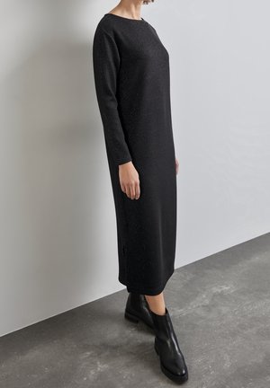 Osoba oblečená v černých dlouhých šatech s dlouhým rukávem a jednoduchým kulatým výstřihem, s černými kotníkovými botami, stojící na šedé podlaze.