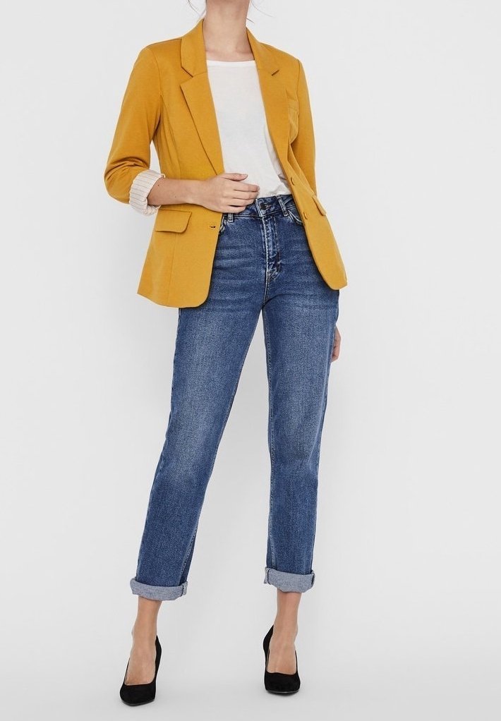 Femme portant un blazer moutarde sur une chemise blanche avec un jean bleu retroussé et des escarpins noirs, debout devant un fond blanc.