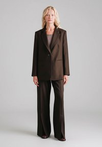 Brązowa marynarka w kroju na miarę z pojedynczym guzikiem, w zestawie z pasującymi szerokimi spodniami z rozcięciami. Gładka tkanina, minimalne detale.