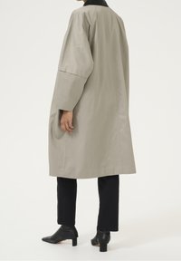 Lang, lichtgrijs jasje met brede mouwen en een zwarte kraag. Draagt over zwarte broeken en spitse zwarte enkellaarzen. Geverfd materiaal.