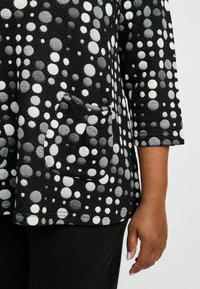 Mustvalge polka dot top, millel on kolmekvart pikkusega varrukad, tekstureeritud kangas ja eesmine tasku. Kujunduses on erineva suurusega täpid.