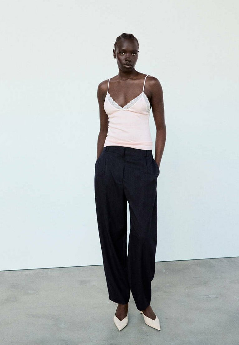 Femme debout, les mains dans les poches, portant un caraco blanc, un pantalon noir ample et des chaussures blanches pointues sur un fond uni.
