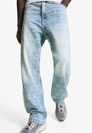 Lichtblauwe spijkerbroek met een patroon van logo's over de hele stof, relaxte pasvorm, rechte pijpen en subtiele vervaging. In combinatie met grijze sportschoenen.
