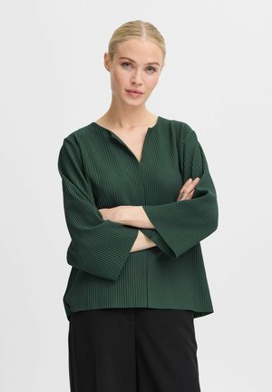 Dark green pleated blouse with a v-neck and three-quarter sleeves, featuring a straight hem and textured fabric. Paired with black pants.