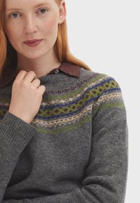 Pull en maille gris avec un empiècement à motif en vert, bleu et bordeaux. Il présente un col rond et des poignets côtelés, superposé sur une chemise à col.