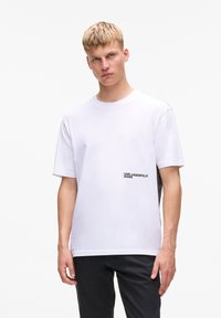 Weißes Baumwoll-T-Shirt mit rundem Ausschnitt, kurzen Ärmeln und einem schwarzen Logo auf der linken Brust. Kombiniert mit dunklen Jeans.