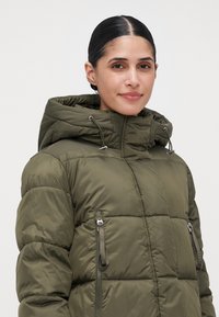 Mujer con cabello oscuro recogido en un moño bajo, vistiendo una chaqueta acolchada de color verde oliva con capucha y bolsillos con cremallera, sonriendo ligeramente.